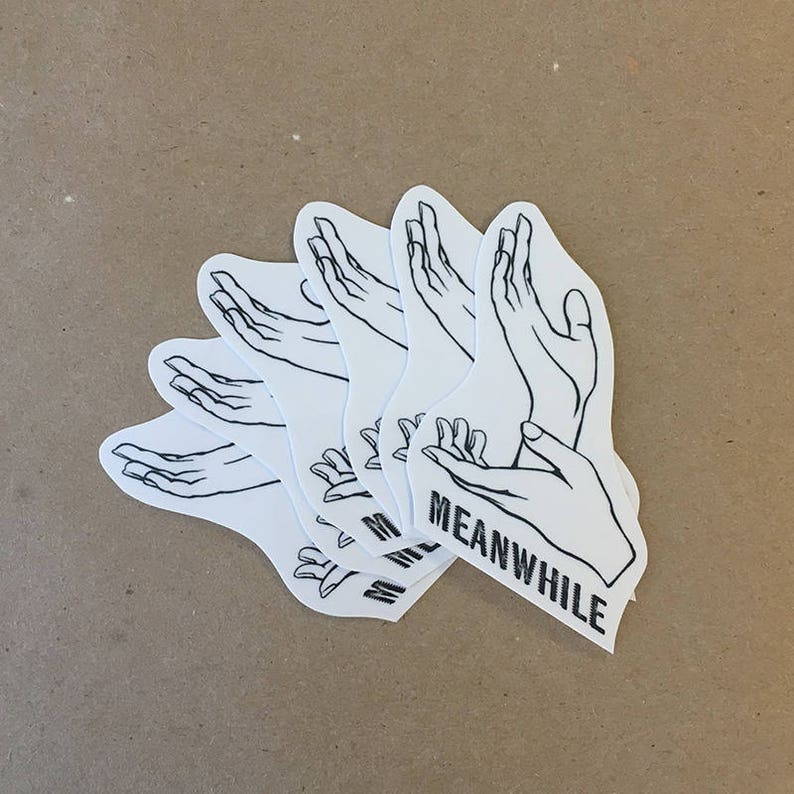 May include: A set of five white stickers with black line drawings of hands. The stickers are layered on top of each other and the top sticker has the text "MEANWHILE" printed on it.