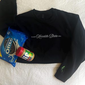 Op de afbeelding: Zwart sweatshirt met de tekst "IN MY Meredith Blake ERA" in witte letters. Het sweatshirt heeft een groene hagedis geborduurd op de mouw. Een pak Oreo-koekjes en een pot pindakaas liggen op een wit oppervlak.
