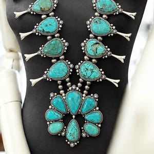 May include: A squash blossom necklace featuring multiple teardrop-shaped turquoise stones set in silver. The design includes a central naja pendant surrounded by smaller turquoise stones, all connected by silver beads and silver blossom accents.