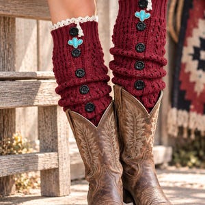 May include: A pair of burgundy knit leg warmers featuring a vertical row of black buttons, a turquoise-colored accent piece, and a white lace trim at the top, worn over brown leather cowboy boots with intricate stitching details on the shaft.