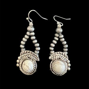 May include: A pair of silver earrings with a teardrop design. Each earring has a white stone in a decorative frame with leaf accents, and a beaded strand. The earrings hang from a silver hook.