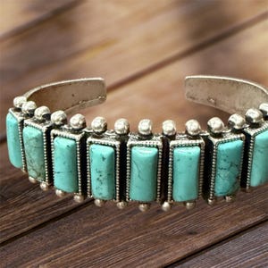 May include: Silver-toned cuff bracelet featuring rectangular turquoise stones. The turquoise stones are set in a silver metal frame and are arranged in a row around the bracelet. The bracelet has a rustic, bohemian style.