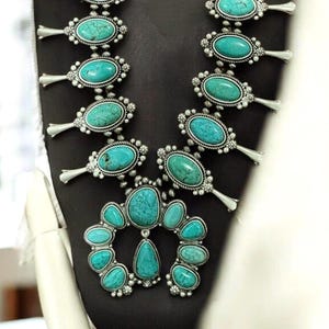 Puede incluir: Un collar llamativo plateado con grandes piedras turquesas ovaladas. El collar tiene un diseño tipo babero con múltiples piedras turquesas y un engaste plateado detallado. El collar se exhibe en un maniquí negro.