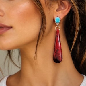 May include: A drop earring featuring a rectangular turquoise stone set in a silver frame, connected by a silver ring to a long, teardrop-shaped red stone with dark black veining and a smooth, polished finish that hangs vertically from the earlobe.