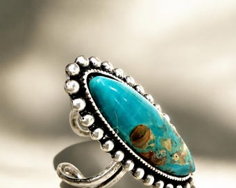 May include: A silver ring featuring a large, oval turquoise stone. The turquoise is a vibrant blue-green with brown and gold inclusions. The ring has a beaded border and a detailed, ornate band.