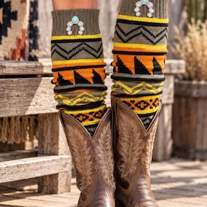 May include: Knit leg warmers in olive green, yellow, orange, and black with a geometric pattern and a silver-toned horseshoe-shaped brooch featuring a central turquoise-colored stone, worn with brown leather cowboy boots with stitched detailing.