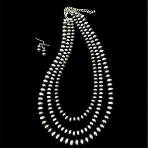Puede incluir: Un conjunto de collar y pendientes plateados sobre un fondo negro. El collar presenta tres hilos de cuentas redondas graduadas. Los pendientes a juego tienen un diseño de cuentas similar. La joyería es probablemente de metal y es adecuada para uso formal o informal.