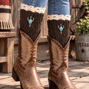 May include: Brown leather cowboy boots with intricate stitching, paired with dark brown knit boot cuffs featuring a cream-colored lace trim at the top and a silver-toned cactus emblem with a central blue oval stone on each cuff.