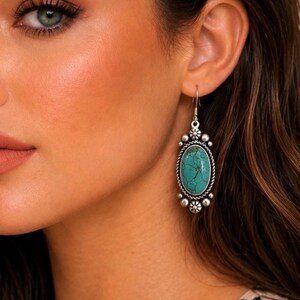 May include: An oval-shaped dangle earring featuring a central turquoise stone with dark veining, encased in a silver-toned metal frame with twisted rope detailing and small decorative silver spheres at the top and bottom of the setting.