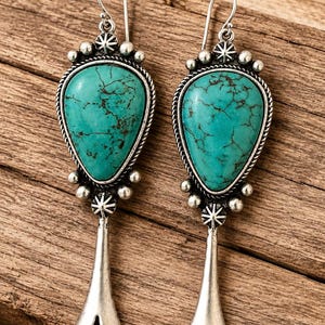 May include: A pair of silver drop earrings featuring a teardrop-shaped turquoise stone with dark veining. The setting includes decorative silver beads, a twisted rope border, and a starburst accent, finished with a long, flared silver base.