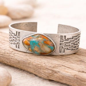 May include: Silver-tone cuff bracelet with a large, oval-shaped stone. The stone features a mix of turquoise, orange, and brown colors. The bracelet has a geometric pattern on the sides, adding a decorative element. Jewelry.