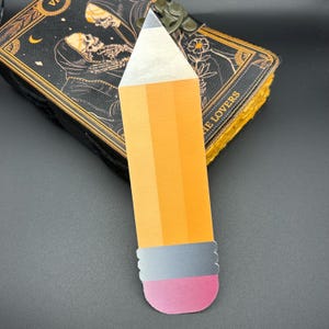 May include: A yellow pencil bookmark with a silver band and a pink eraser. The bookmark is laying on top of a closed book with a black cover and gold accents.