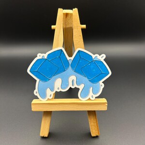 May include: Two blue ice cubes melting and dripping on a white background. The ice cubes are in the shape of a square with rounded corners. The dripping water is a light blue color.