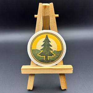 May include: A round sticker featuring a green pine tree silhouette against a yellow sun and green mountains. The design is set within a yellow and green border. The sticker is displayed on a small wooden easel against a dark background.