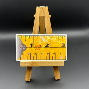 May include: A yellow ruler with a holographic finish. The ruler measures 3 inches and has markings for both inches and centimeters. The ruler is on a small wooden easel.