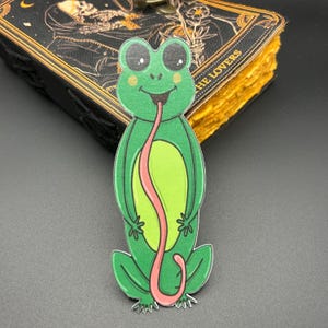May include: A green frog with a long pink tongue sticking out of its mouth. The frog is cartoon-style and has big eyes. The frog is standing on its hind legs.