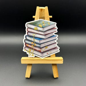 May include: A stack of six vintage books with a vine growing up the side. The books are various shades of blue, green, red, and brown. The books are on a small wooden easel.