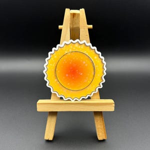 May include: A white sticker with a yellow and orange sun design. The sun has a white outline and white stars inside. The sticker is on a small wooden easel.