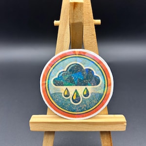 May include: A round sticker featuring a stylized cloud with raindrops. The cloud is dark blue and green, with yellow raindrops. The sticker has a teal and orange border and is displayed on a small wooden easel.