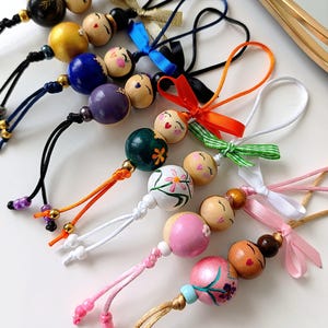 Può includere: Una collezione di ornamenti di perline di legno dipinte a mano. Ogni ornamento presenta un volto dipinto ed è impreziosito da nastri e cordoncini in vari colori, tra cui nero, blu, arancione e rosa. Le perline sono dipinte in una varietà di colori e disegni.