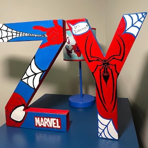 Peut inclure: Lettres en bois "ZY" rouges, bleues et blanches, avec des motifs Spider-Man. La lettre "Z" présente une toile et une main, et la lettre "Y" un emblème d'araignée. Un logo Marvel est sur la base.