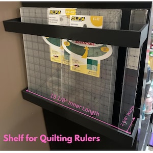 Puede incluir: Estante negro diseñado para reglas de acolchado. El estante tiene una longitud interior de 38,4 cm y un ancho de 6,7 cm. Contiene reglas de acolchado transparentes, incluidas reglas de la marca OLFA.