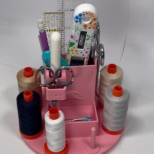 Könnte beinhalten: Ein rosa Nähkörbchen-Karussell-Turm mit Garnrollen, Scheren und anderem Nähzubehör. Das Körbchen hat eine runde Basis und ein zentrales Fach für Werkzeuge. Das Bild enthält den Text "Ray of Sonshine Design" und "Sewing Caddy Carousel Tower."