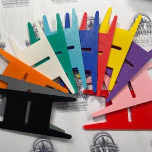 Puede incluir: Una colección de bobinas de hilo planas y multicolores dispuestas en forma de abanico. Las bobinas están hechas de plástico sólido y cuentan con una ranura rectangular central con dos puntas en cada extremo. Los colores incluyen negro, gris, naranja, blanco, verde, turquesa, azul, rojo, amarillo, morado y rosa.