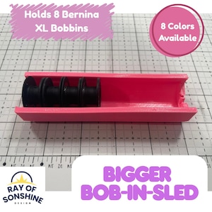 May include: A bright pink plastic bobbin holder labeled "Holds 8 Bernina XL Bobbins" and "8 Colors Available" containing four black sewing machine bobbins. The text "BIGGER BOB-IN-SLED" and the logo "RAY OF SONSHINE DESIGN" are visible in the image.