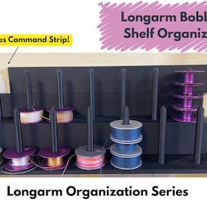 May include: A black shelf organizer for longarm quilting bobbins. The organizer has multiple vertical rods to hold bobbins and a shelf for storing additional bobbins. The text "Longarm Bobbin Shelf Organizer" is written in pink on a pink background. The text "Hides Command Strip!" is written in yellow on a yellow background.