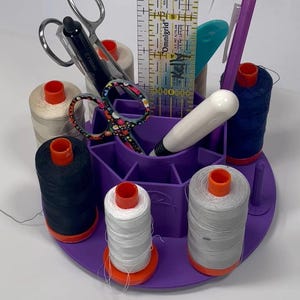 May include: A purple sewing caddy carousel containing spools of thread, scissors, a ruler, and a pencil. The caddy has multiple compartments for organising sewing supplies. The text "Sewing Caddy Carousel" is displayed on the image.