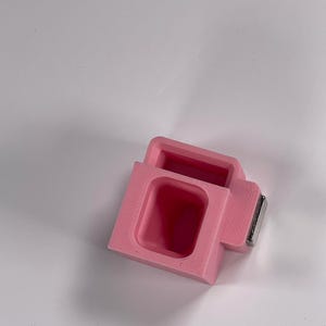 May include: A pink, rectangular object with a recessed interior and a small, rectangular metal piece attached. The object has a boxy shape with a rectangular opening. The item is likely a small container or holder.
