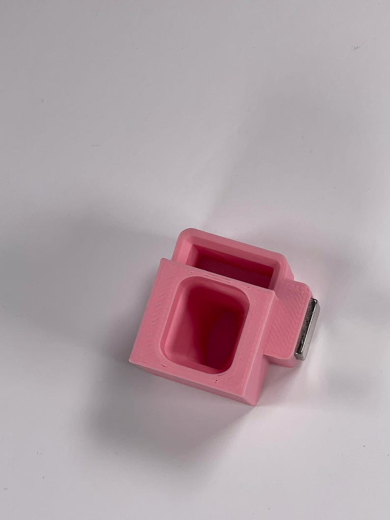 May include: A pink, rectangular object with a recessed interior and a small, rectangular metal piece attached. The object has a boxy shape with a rectangular opening. The item is likely a small container or holder.