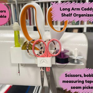May include: White plastic long arm caddy shelf organiser with a handle. It holds a pair of pink and black scissors, a pair of orange and white scissors, a seam ripper, a measuring tape, two bobbins, and a seam picker. The text "Long Arm Caddy Shelf Organiser" is written on a pink background.