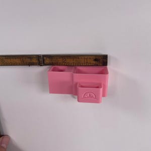 May include: A pink plastic desk organizer with three compartments and a small drawer. The organizer is approximately 3 inches wide and 2 inches tall. The drawer has a curved design on the front.