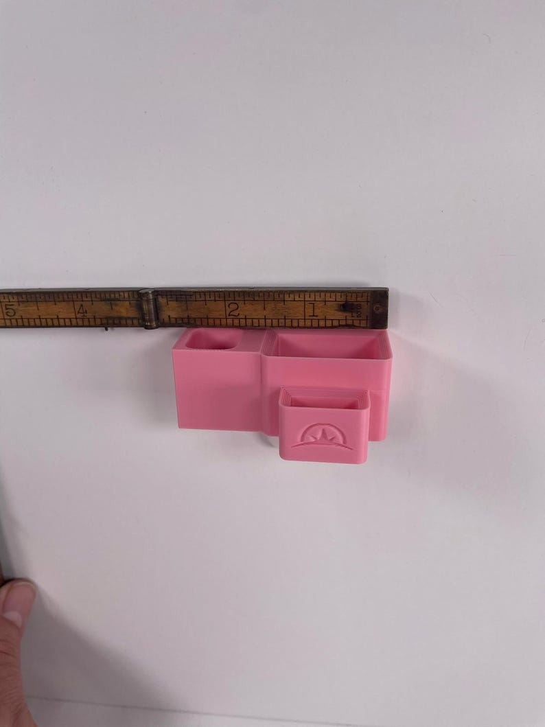 May include: A pink plastic desk organizer with three compartments and a small drawer. The organizer is approximately 3 inches wide and 2 inches tall. The drawer has a curved design on the front.