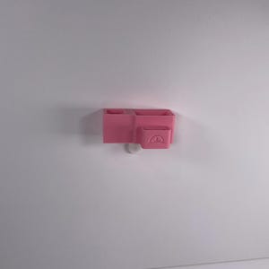 May include: A pink, wall-mounted organizer with multiple compartments. The organizer has two rectangular sections and a smaller, square section with a curved design. The organizer is made of plastic and is mounted on a white wall.