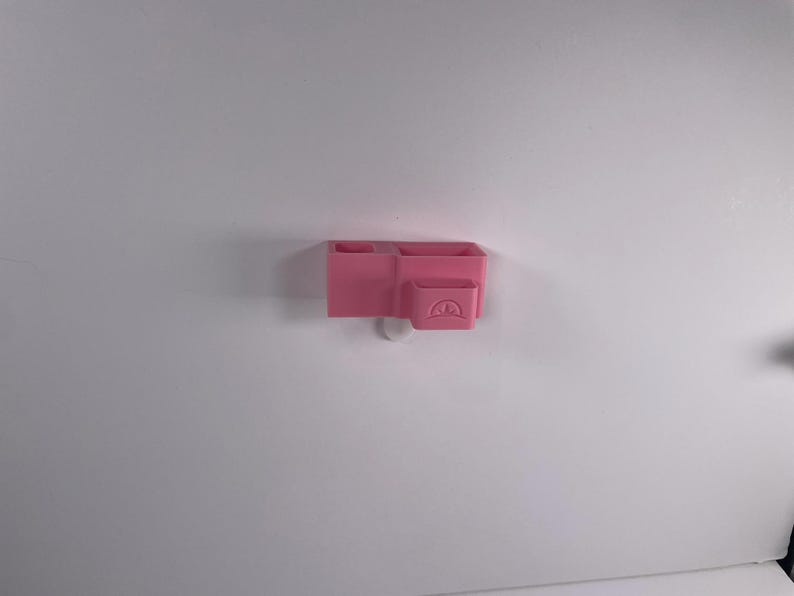 May include: A pink, wall-mounted organizer with multiple compartments. The organizer has two rectangular sections and a smaller, square section with a curved design. The organizer is made of plastic and is mounted on a white wall.