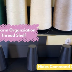 May include: A black thread shelf with three spools of thread, one green, one white, and one beige. The shelf is labeled "Longarm Organization Thread Shelf" and is mounted on a wall using Command Strips, which are hidden behind the shelf.