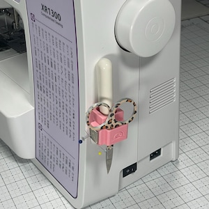 May include: A white sewing machine with a pink and leopard print scissors holder. The scissors have a leopard print design. The machine has the text "XR1300 Computerized Sewing".