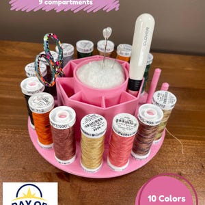 Op de afbeelding: Roze draaiende knutselwagen met 16 spoelhouders en 9 vakken. De wagen heeft een wit naaldkussen en een witte Clover naaldinvoerder. De tekst "Hand Work Rotating Caddy with 16 Spool Holders 9 compartments" is in roze op een witte achtergrond geschreven.