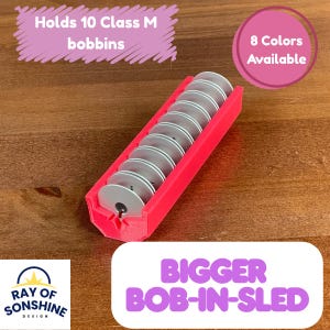 May include: A bright pink bobbin sled holding ten silver bobbins. The text on the image says "Holds 10 Class M bobbins" and "8 Colors Available". The product is labeled "BIGGER BOB-IN-SLED" with the Ray of Sonshine Design logo.
