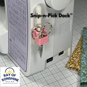 May include: A pink Snip-n-Pick Dock™ attached to a sewing machine, holding scissors. The scissors have a leopard print design. The sewing machine is white. The text "Snip-n-Pick Dock™" is visible.