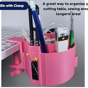 May include: A pink handy bin with clamp, designed for organizing sewing or craft supplies. The bin holds various tools, including scissors, pens, and a measuring tape. The text "Handy Bin with Clamp" is visible on the top left.