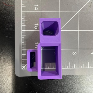 May include: A purple, 3D-printed object with a rectangular shape and a hollow interior. The object is positioned on a gray cutting mat with measurement markings. The object's dimensions appear to be approximately 1.5 inches in height.