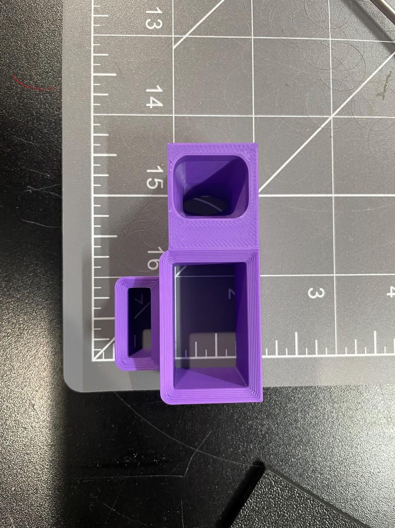 May include: A purple, 3D-printed object with a rectangular shape and a hollow interior. The object is positioned on a gray cutting mat with measurement markings. The object's dimensions appear to be approximately 1.5 inches in height.