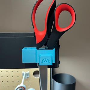 May include: A pair of scissors with black handles and red finger holes is held in a light blue 3D-printed holder. The holder is mounted on a black metal surface. The scissors are partially visible, with the blades extending downwards.