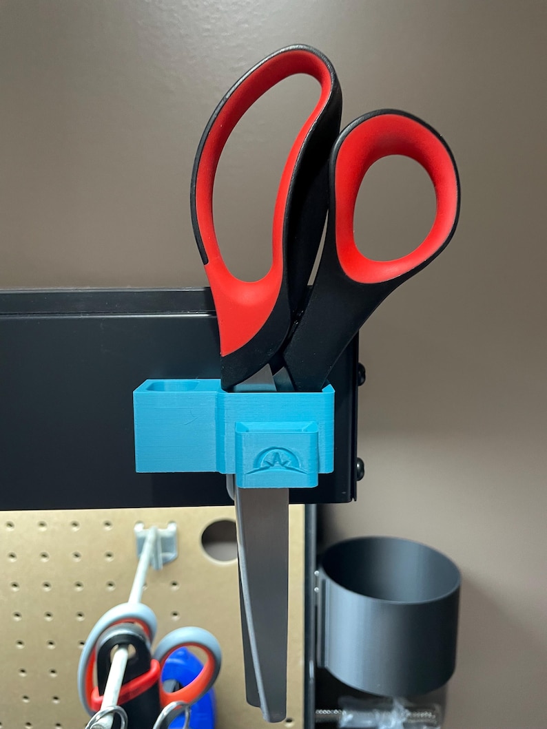 May include: A pair of scissors with black handles and red finger holes is held in a light blue 3D-printed holder. The holder is mounted on a black metal surface. The scissors are partially visible, with the blades extending downwards.