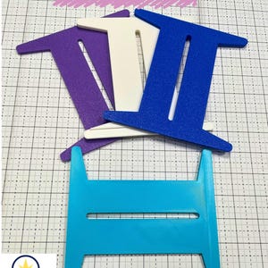 Pode incluir: Uma coleção de Binding Spools XL coloridos em roxo, branco, azul e turquesa. As bobinas estão dispostas sobre uma superfície quadriculada. A imagem inclui o texto "Binding Spools XL" e o logotipo "Ray of Sunshine Design".