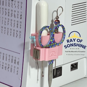 Puede incluir: Un organizador Magnetic Snip-n-Pick Sidecar de plástico rosa claro fijado a una máquina de coser. Contiene una herramienta blanca, unas tijeras con un patrón de huellas de colores y varias agujas de coser sujetas por un imán plateado.